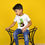 Thumbnail: one more photo of a kid sitting and wearing a t shirt by Hugs&Hues having a dinosaur and number 5 embroidered on it and posing