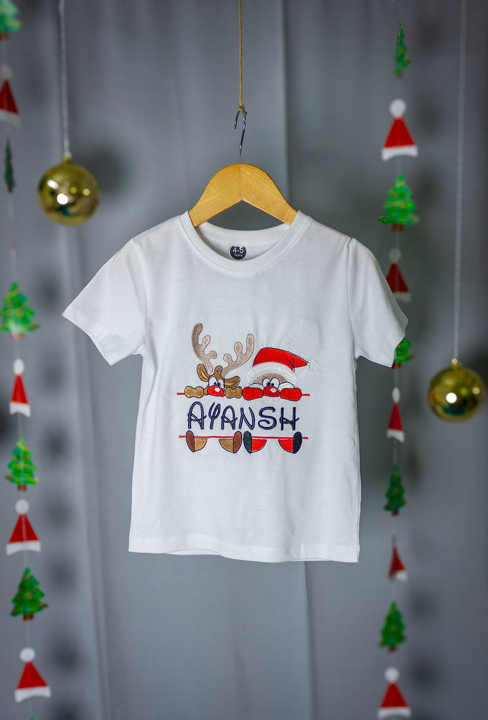 Thumbnail: a t shirt from Hugs&Hues Christmast collection having a santa clause, reindeer and child name embroidered on it and hanging in front of christmas decorations