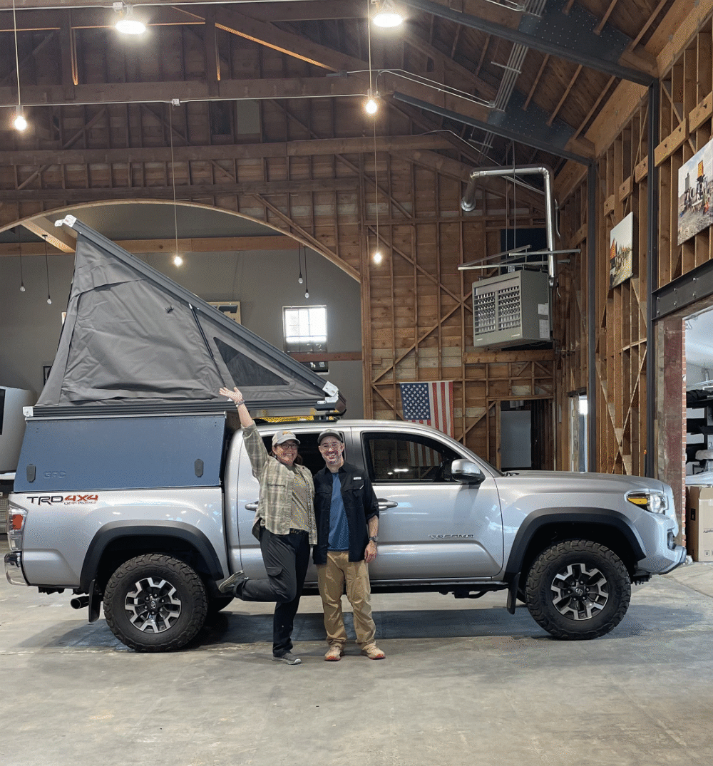 Nikki and Jason posing for a photo in front of their new Go Fast Camper in a large warehouse - via NVilloria.com