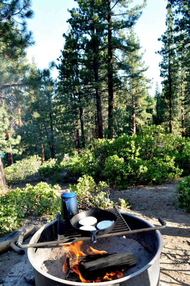 a campfire ring and grill. a pan with pancakes and a blue teapot are sitting on top of the grill.