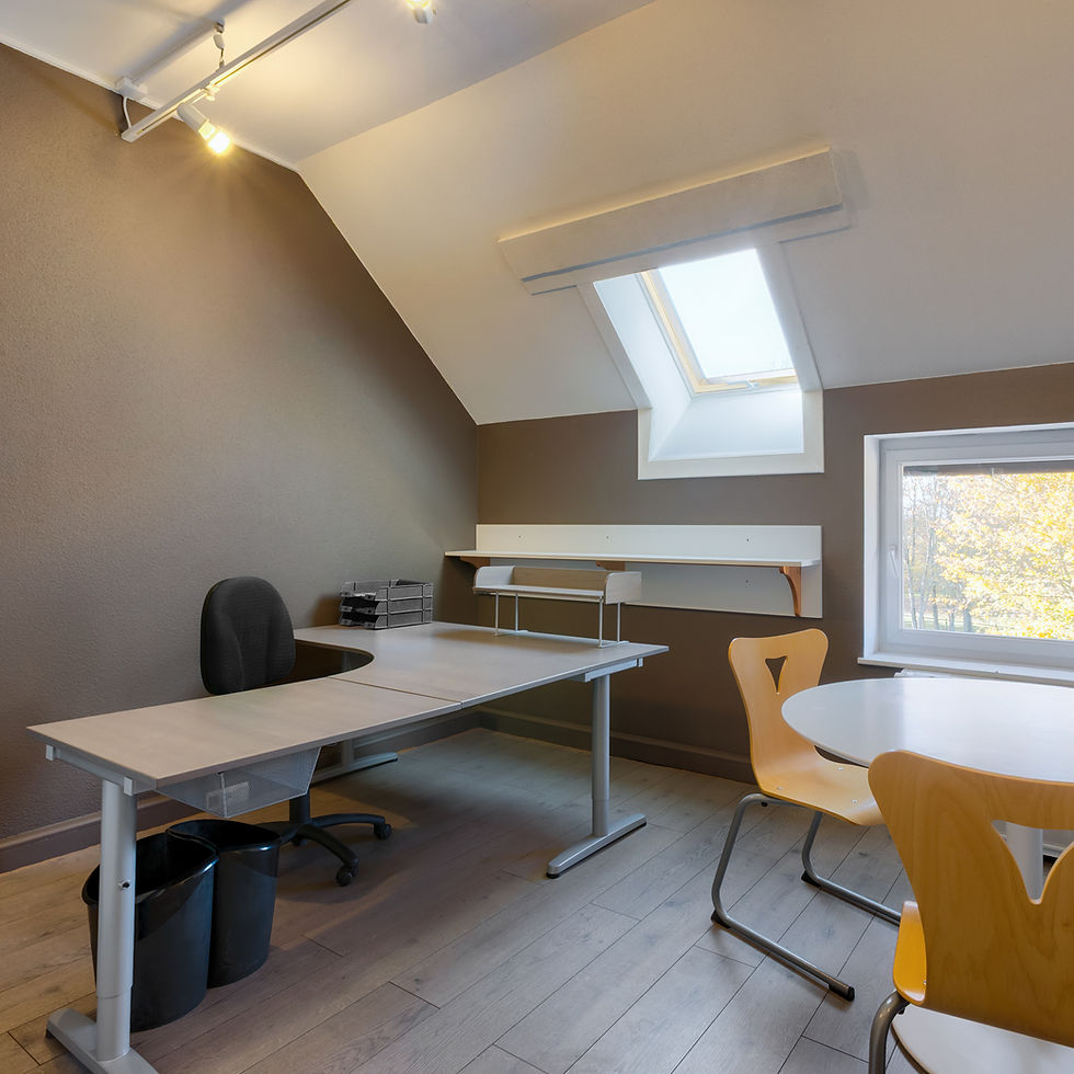 Un bureau avec plafond mansardé, des murs de couleur taupe et 2 fenêtres. En plus du bureau principal il y a une table de réunion