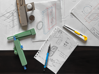 Technical sketches and tools on a dark surface, featuring a yellow utility knife, gears, and a green device. Plans depict mechanical concepts.