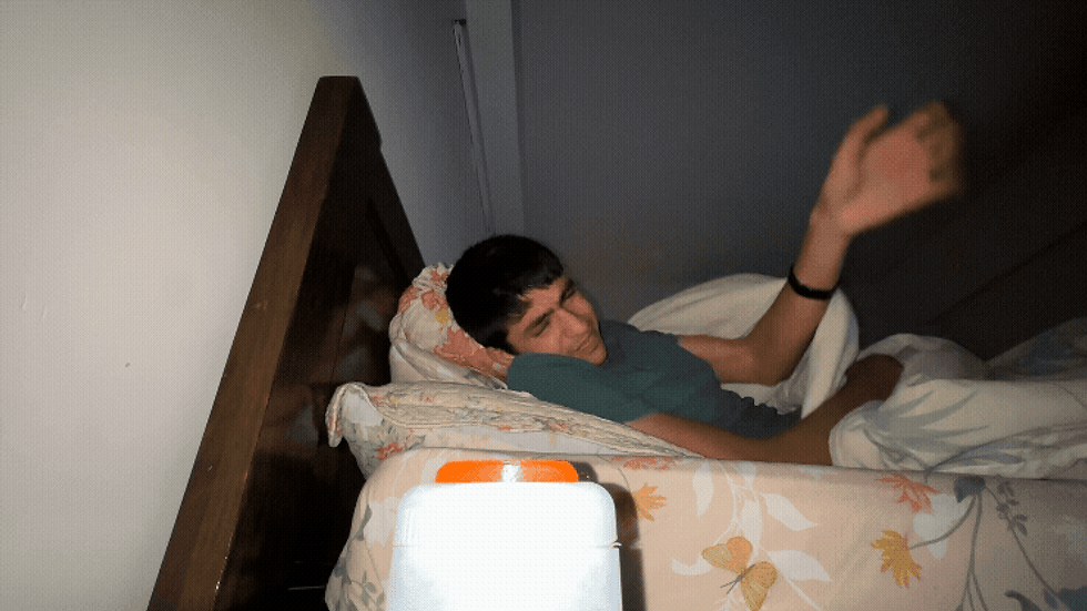 Man in bed, startled, raises hand defensively. Dim room, floral bedspread. Alarm clock with orange details on beside table. Surprised mood.