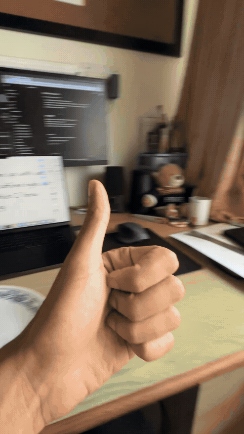 Hand giving thumbs up in a home office with blurry computer screens in the background. Warm lighting and a slight motion blur create a dynamic feel.