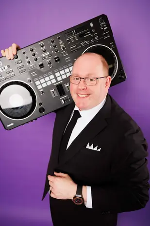 DJ Dan Fudim, NYC DJ, is holding a Pioneer DJ controller on his shoulder while dressed in a black suit with a white pocket square and a black tie, standing against a purple background.