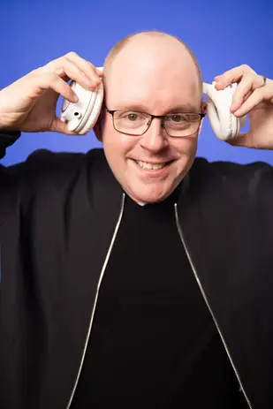 DJ Dan Fudim, NYC DJ, holding white headphones up to both ears against a blue background.