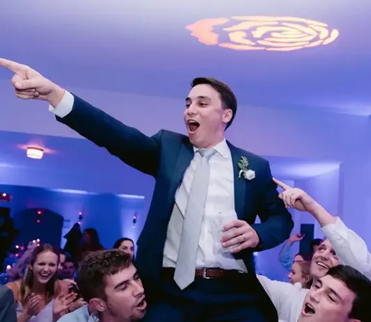 DJ Dan Fudim, NYC DJ, at a lively event with a person in a suit being lifted up by a group of people, pointing upwards with a drink in hand, under a decorative light pattern on the ceiling.