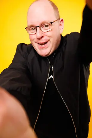 DJ Dan Fudim, NYC DJ, posing against a vibrant yellow background in a black jacket with a zipper