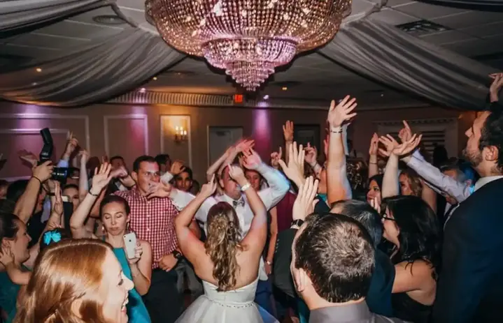 DJ Dan Fudim, NYC DJ, energizing the crowd at a lively event, with people dancing and raising their hands under a grand chandelier in an elegantly decorated venue.