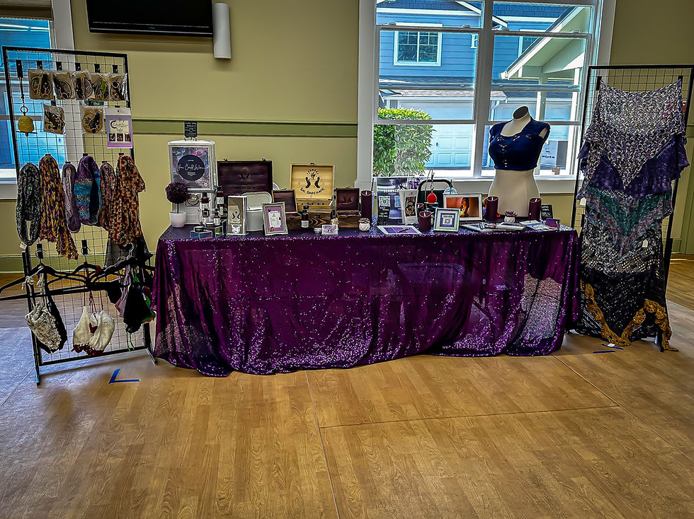 Craft display with mannequin and accessories on a table covered in a purple sequined cloth. Products hang on racks. Bright room with large window.
