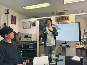 男性がモニターの前に立ち各責任者に事業の進捗を報告している
