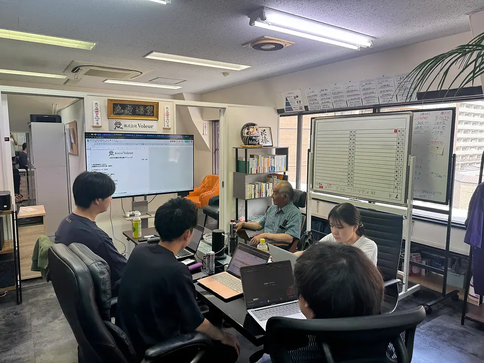 モニターの前で責任者の男性が説明しているのを各事業部の責任者が聞いている。