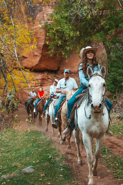6 people ride horseback in a line being led by a cowgirl on a white horse through Cave Lakes private resort canyon.
