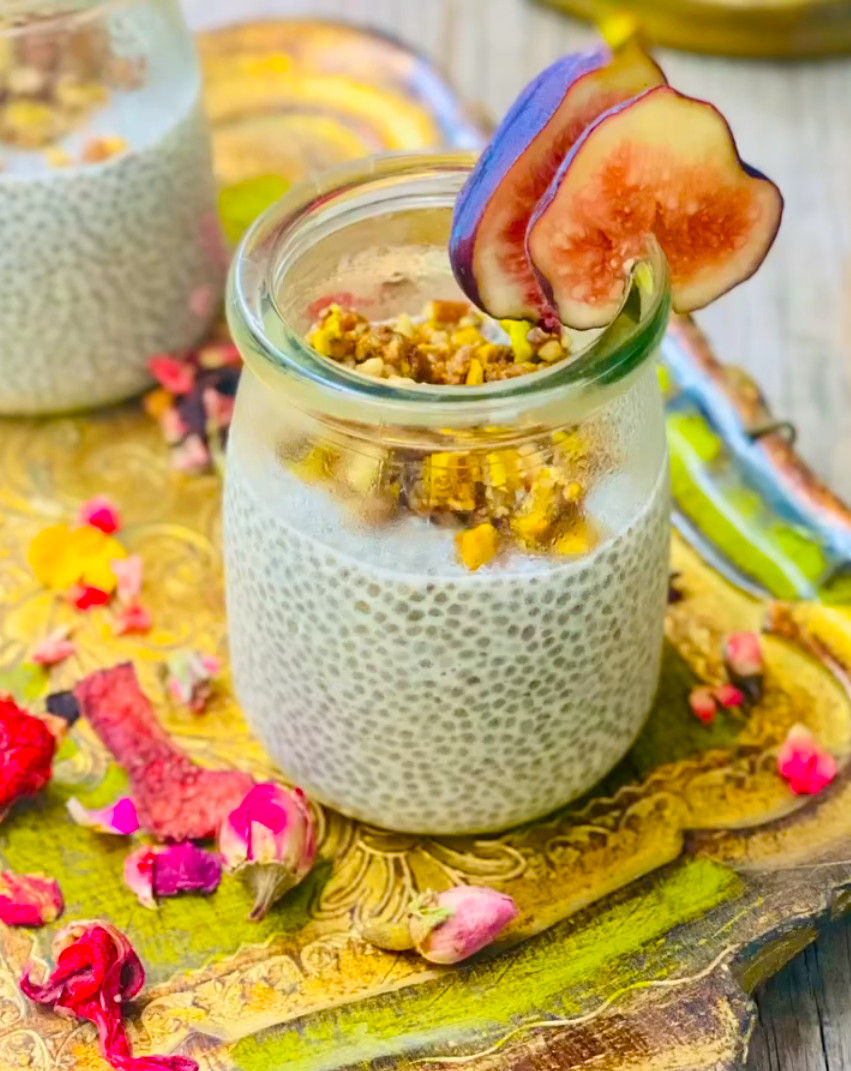 Vasito de pudín de chía con higos y nueces, sobre una bandeja colorida con pétalos; fondo desenfocado, aspecto apetitoso.