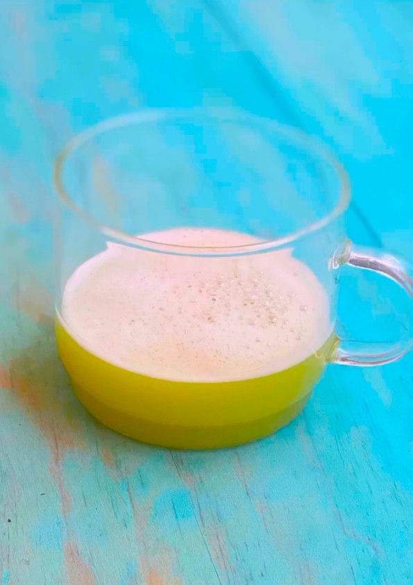 Taza de vidrio con bebida verde con un toque amarillo y espuma blanca sobre mesa de madera turquesa, fondo sereno.
