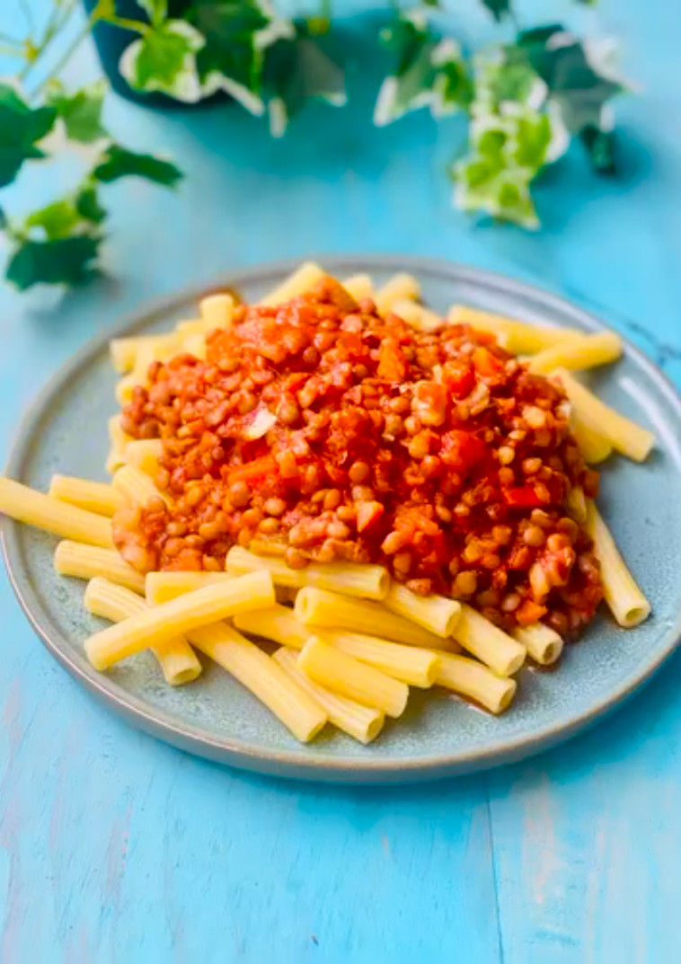 Pasta con salsa de lentejas y tomate en un plato gris sobre mesa azul, con hojas verdes desenfocadas.