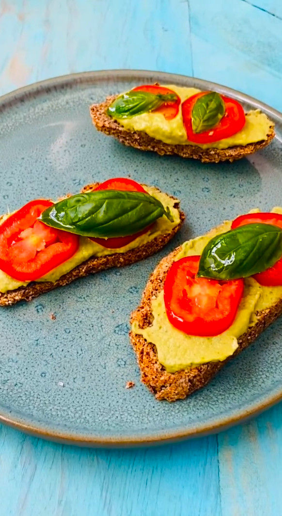 Tres tostadas con crema de aguacate, tomate y albahaca en un plato azul sobre mesa turquesa.