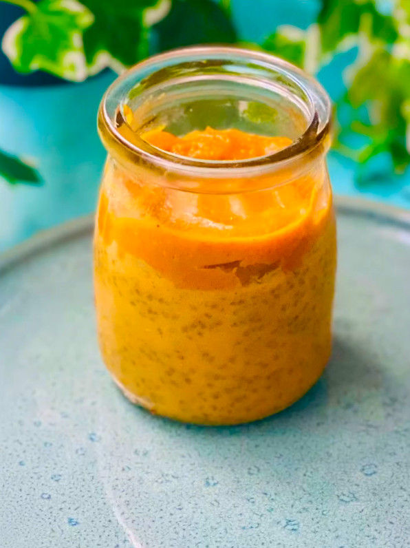 Tarro de vidrio con postre crema naranja sobre mesa turquesa, con hojas verdes desenfocadas al fondo.