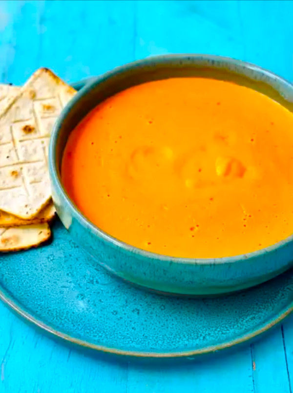 Sopa naranja en un cuenco azul sobre plato turquesa, con galletas saladas al lado, sobre mesa azul.