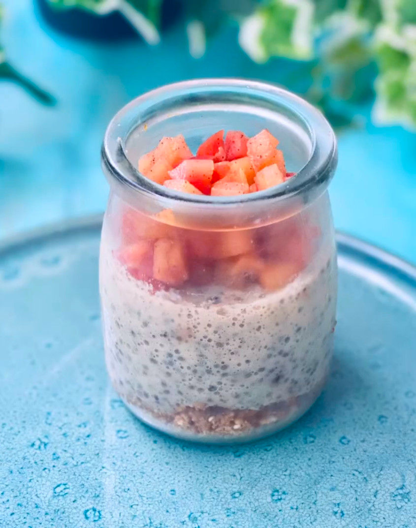 Tarro de pudín de chía con fruta picada encima, sobre mesa azul; fondo desenfocado verde y tono fresco.
