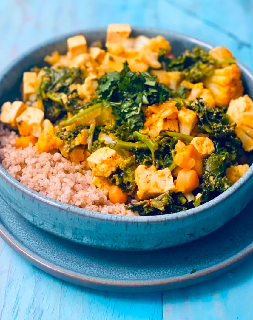 Bol de quinoa con kale, garbanzos y cubos de tofu, sobre mesa azul; plato saludable y colorido.