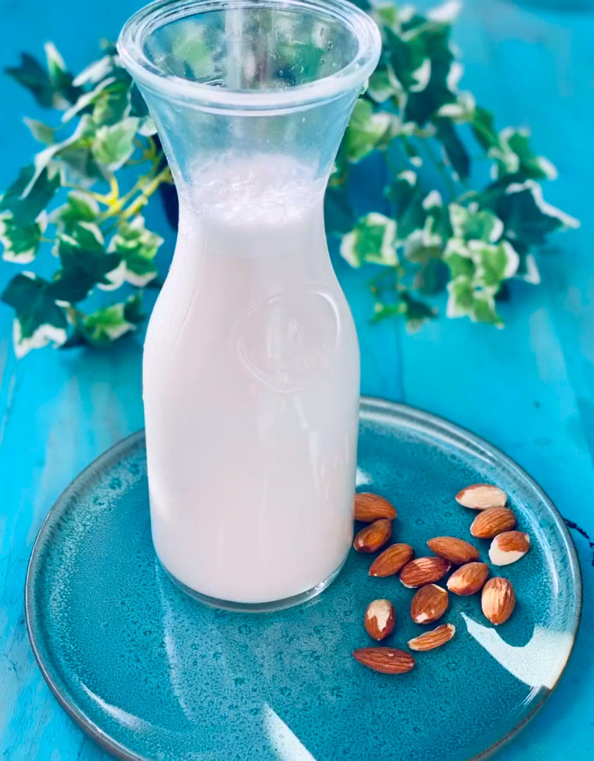 Botella de leche de almendras sobre plato azul, con almendras alrededor y hojas verdes sobre fondo turquesa.