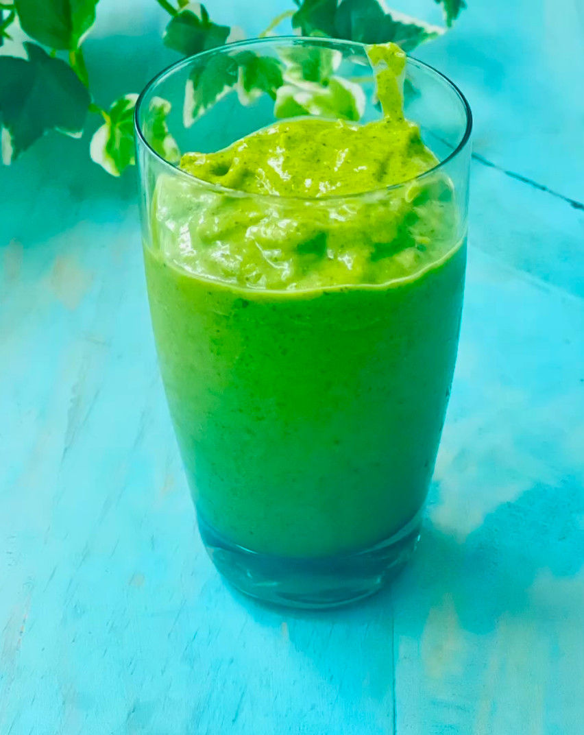 Vaso de batido verde espeso sobre mesa turquesa, con hojas desenfocadas al fondo; aspecto fresco y natural.