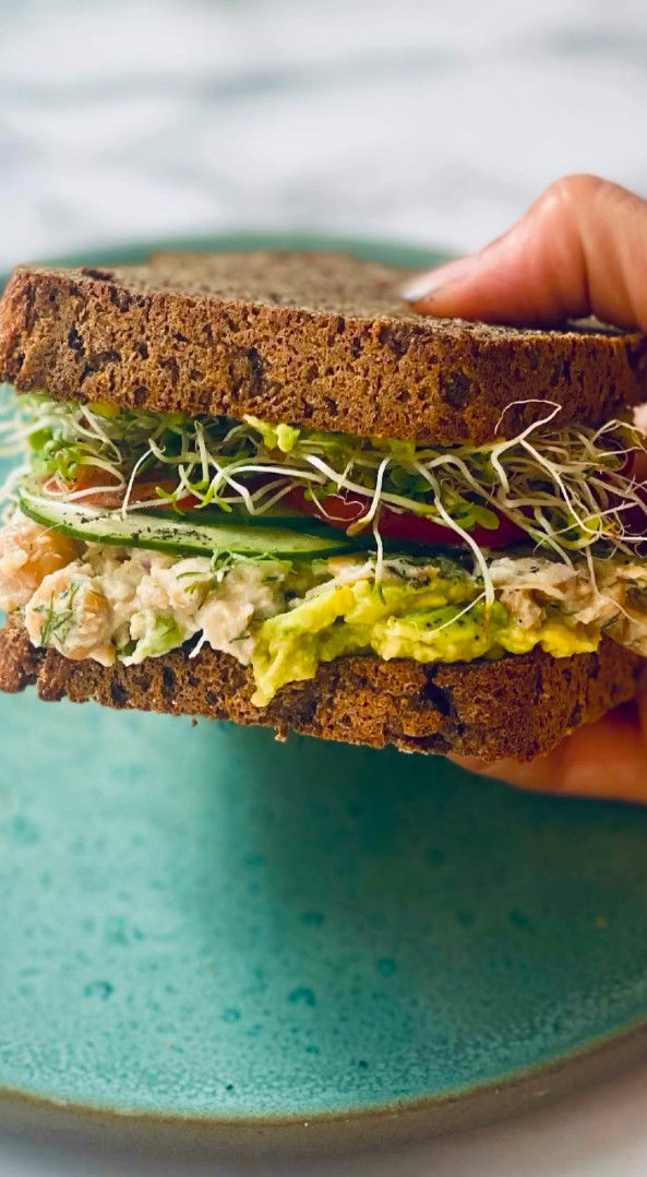 Mano sostiene un sándwich de pan oscuro con brotes, pepino y aguacate sobre un plato turquesa.