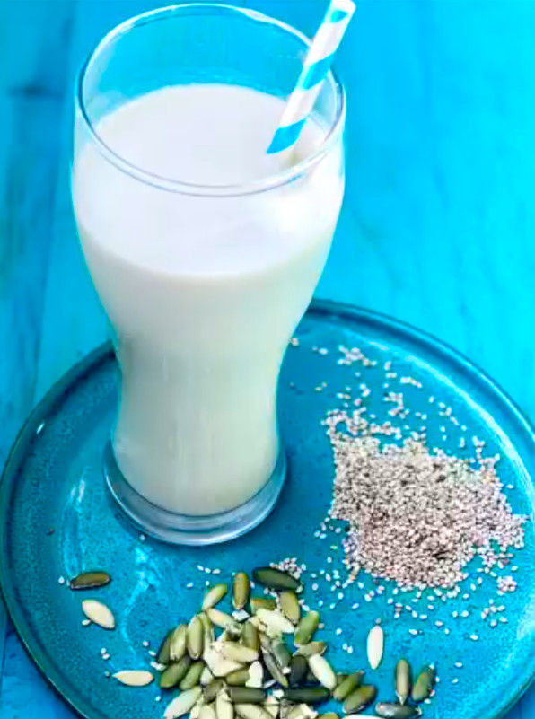 Vaso de leche con pajita azul sobre plato azul, rodeado de semillas y granos; ambiente fresco.