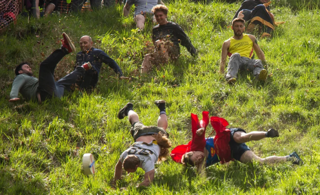 Several people running and falling down a slope of grass and mud in Gloucestershire, England, for the Cooper's Hill Cheese-Rolling Festival.