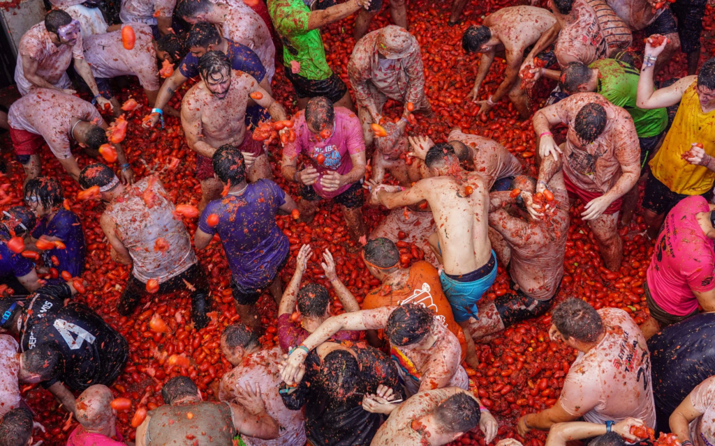 Crowd of people covered in smashed tomatoes participating in La Tomatina, a large tomato-throwing festival in Spain, surrounded by a sea of red pulp and flying tomatoes.