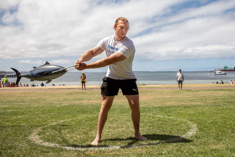 Man participating in a tuna throwing competition on a grassy field near the beach, holding a fake fish by a string mid-swing, with spectators and the ocean in the background.