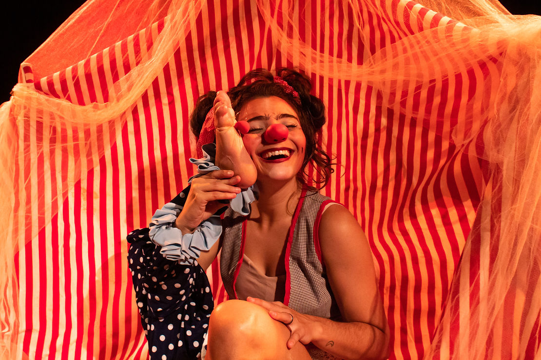 A atriz Emeli Bruna vestida de palhaça segura seu pé com nariz de palhaço próximo ao rosto.