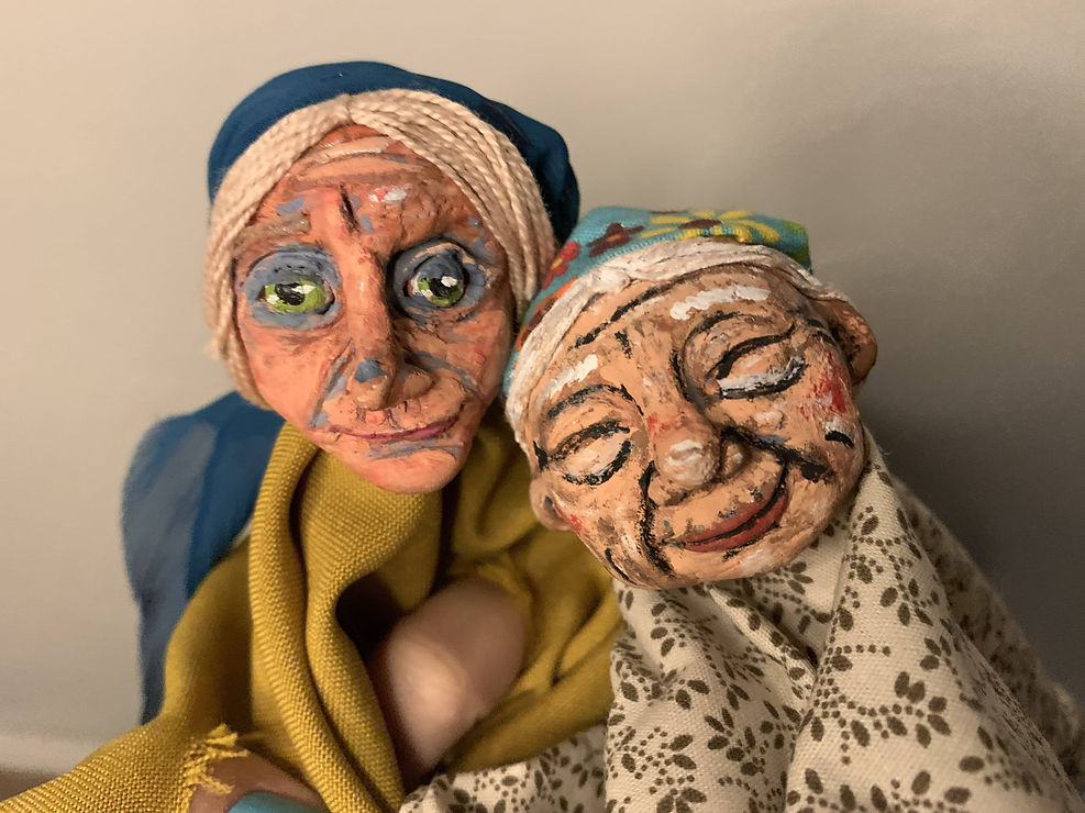 As mãos da atriz Amália Leal se tornam o corpo de uma bruxa de pele e cabelos brancos, lenço azul na cabeça e roupa verde. Abraçada à ela está uma outra figura de pele e cabelos brancos com o lenço de cabeça e roupa floridas.