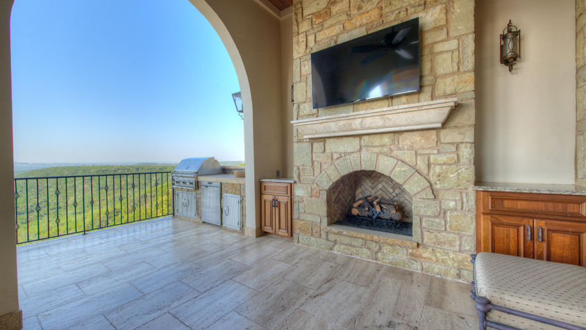 Outdoor kitchen with stone fireplace and a mounted TV, overlooking scenery.