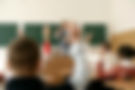 Students raising hands in classroom with teacher present at blackboard.