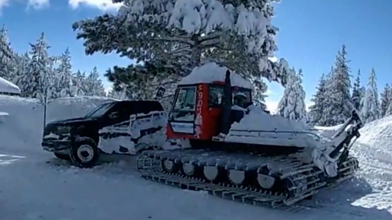 Red snowplow clearing snow next to a black truck in a snowy forest with pine trees under a clear blue sky. Snow covers the ground and vehicles.