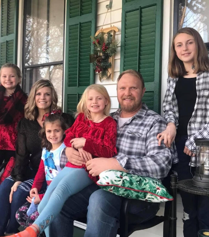 Stephanie Barton with her husband Bill and their four daughters. The family is sitting on a porch posing together and smiling in front of a white house with green shutters.