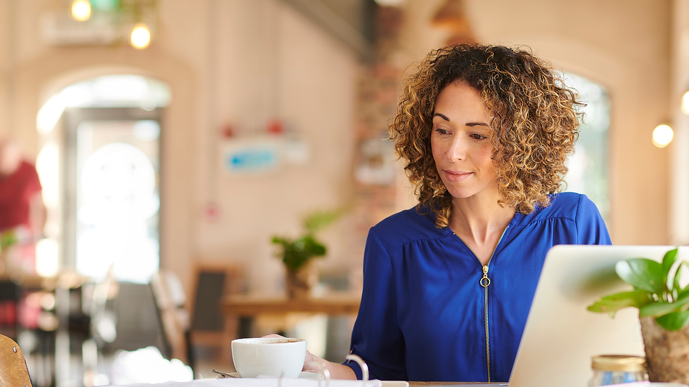 Woman in a blue top sits in a cafe, looking at a laptop with a coffee cup beside her. Bright, cozy setting with blurred background.