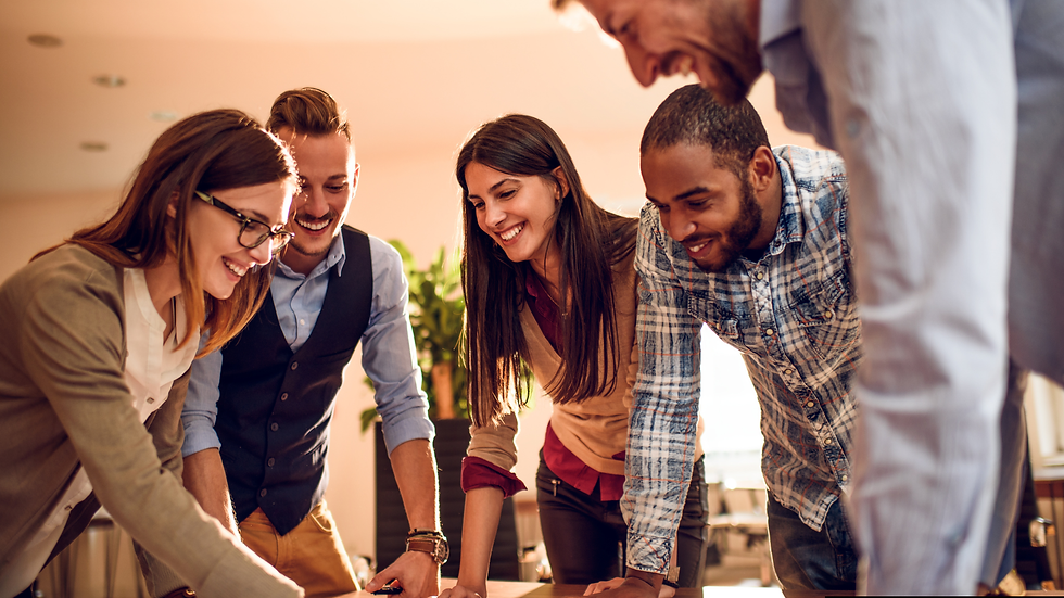 Five people smiling, leaning over a table, collaborating in a bright office. Casual attire. Warm light enhances a positive, focused mood.