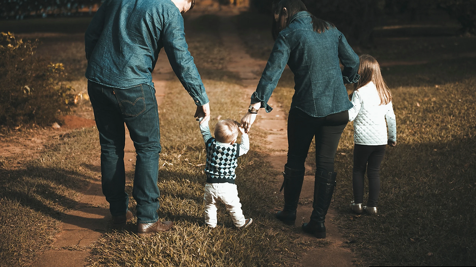 Family of four holding hands, walking away on a dimly lit dirt path at night, wearing denim and dark clothes.