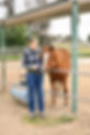 Smiling woman in plaid shirt petting a brown horse at the paddock.