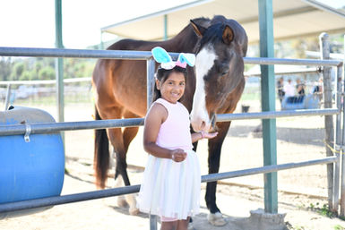 Smiling girl wearing bunny ears with brown horse behind fence.