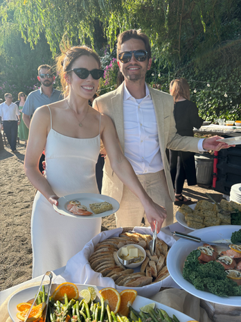 Smiling woman and man serve food at an elegant outdoor buffet.