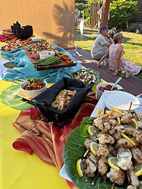 A long table with colorful table clothes full of bite sized appetizers outside in front of a couple sitting down and laughing. 