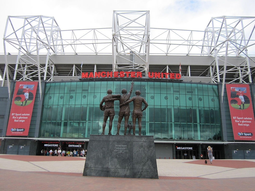 Statue of three footballers outside Manchester United stadium. Red signage, banners, glass facade. Overcast sky creates a calm mood. Old Trafford,