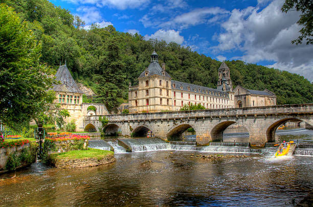 Brantôme Abbey and its stone bridge over the River Dronne, with a canoeist passing beneath a bright blue sky.