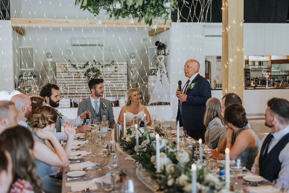 Father giving wedding speech at long reception table with bride, groom, candles and greenery décor under fairy lights.