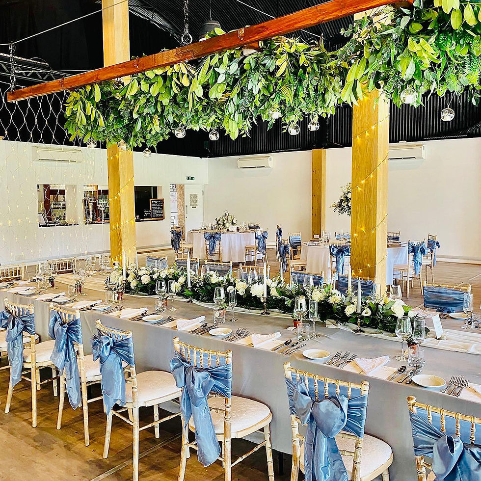 Wedding banquet hall with long tables, blue chair sashes, greenery ceiling installation and white floral garlands.