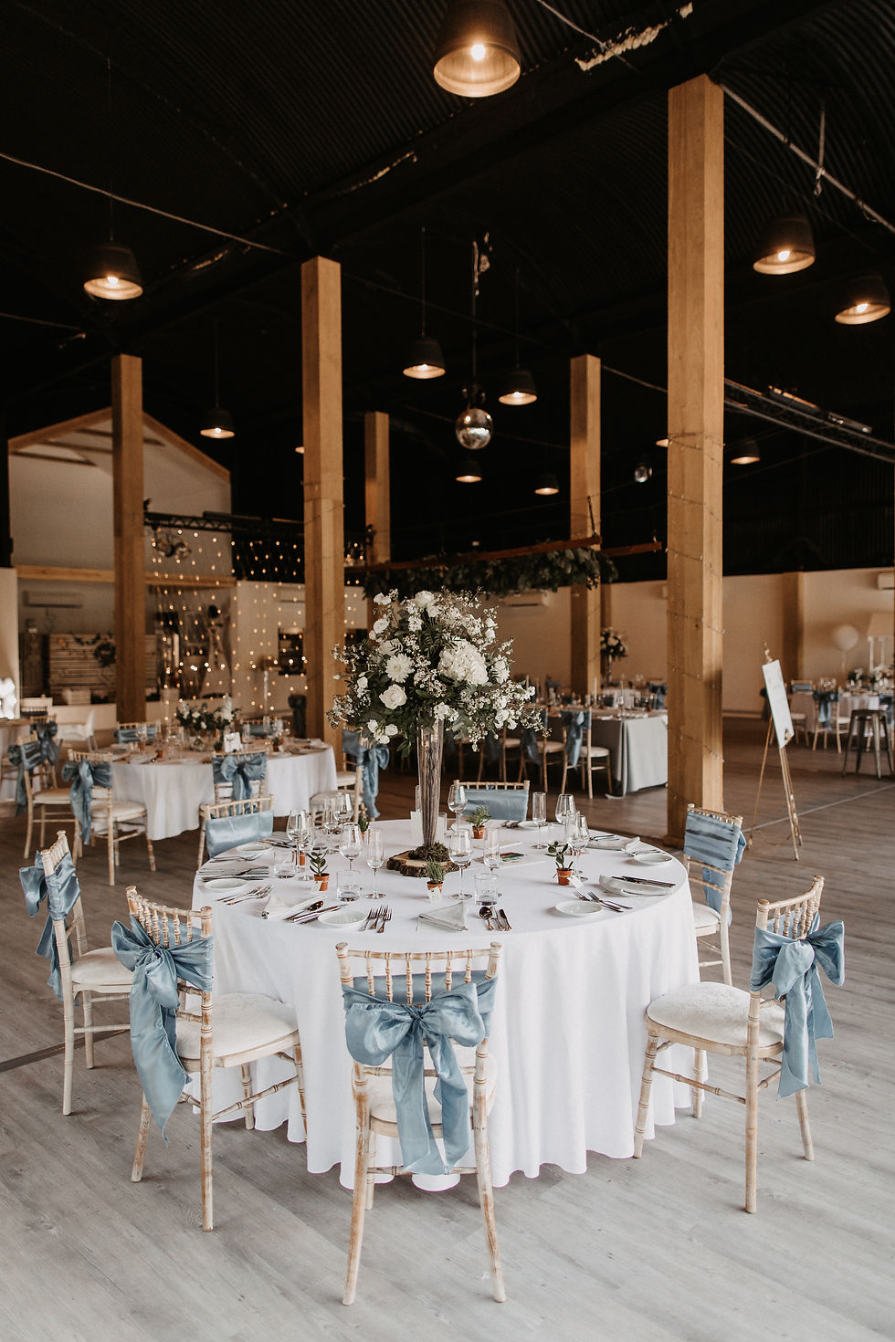 Wedding reception setup with round tables, tall white floral centrepieces and blue chair sashes in a rustic barn venue.